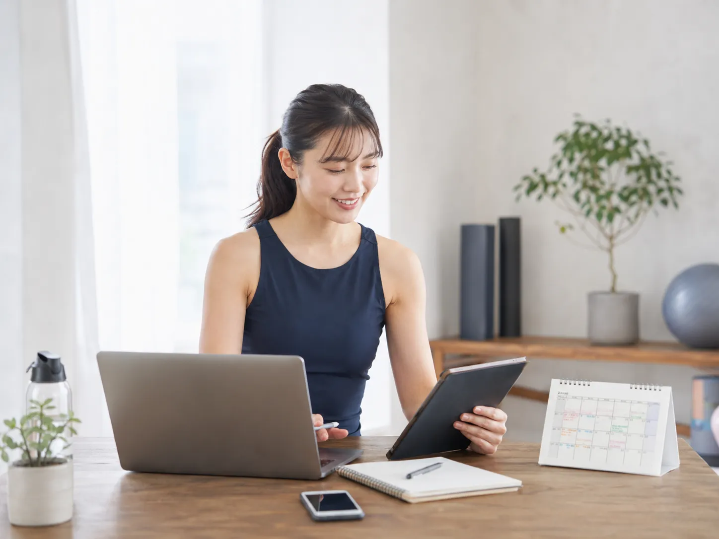 女性パーソナルトレーナーがパソコンとタブレットで予約や顧客情報を管理している様子